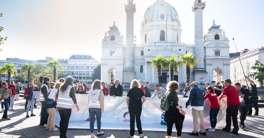 Langer Tag der Flucht Karlsplatz  Eine Menschengruppe hält ein großes Transparent in den Händen und steht vor der Karlskirche